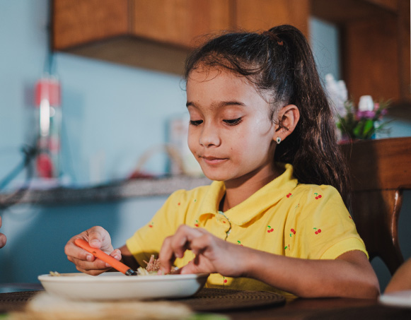 A young girl in a yellow shirt is eating while sitting at a table.
