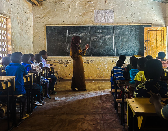 Shadows cast a long classroom, where children are focused on a teacher at the chalkboard.
